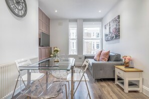 Uma sala de estar moderna com uma mesa de jantar de vidro, cadeiras brancas, um sofá cinza e uma pequena mesa de apoio de madeira com um vaso.