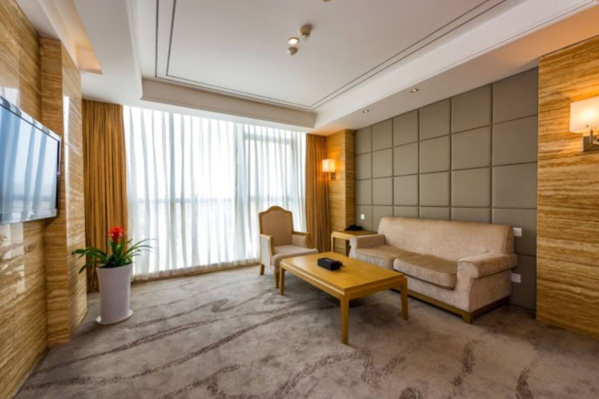 Ein modernes Hotelzimmer mit großem Fenster, einem Flachbildfernseher, einem beigefarbenen Sofa, einem Holztisch und einem Topfpflanzen.