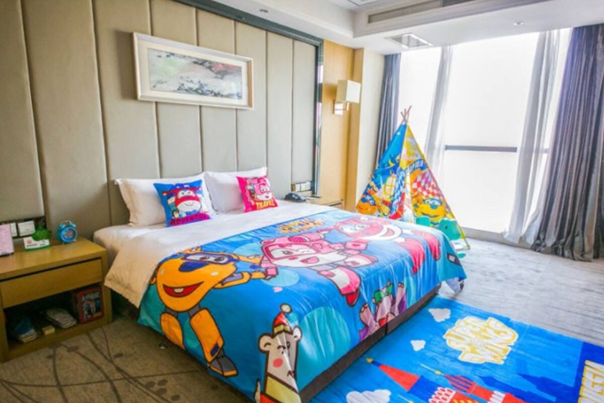 Ein Hotelzimmer mit einem Bett, das eine farbenfrohe Bettdecke mit Zeichentrickmotiven, einen Nachttisch mit einer Uhr, ein gerahmtes Bild an der Wand und ein Fenster mit Vorhängen zeigt.