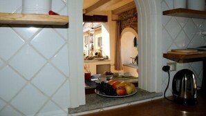 Une cuisine avec un mur recouvert de carreaux, des étagères en bois, une bouilloire et un plat de fruits.