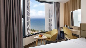 Habitación de hotel con una ventana grande que ofrece vistas al océano, una cama, un escritorio con silla y una pequeña mesa con fruta.