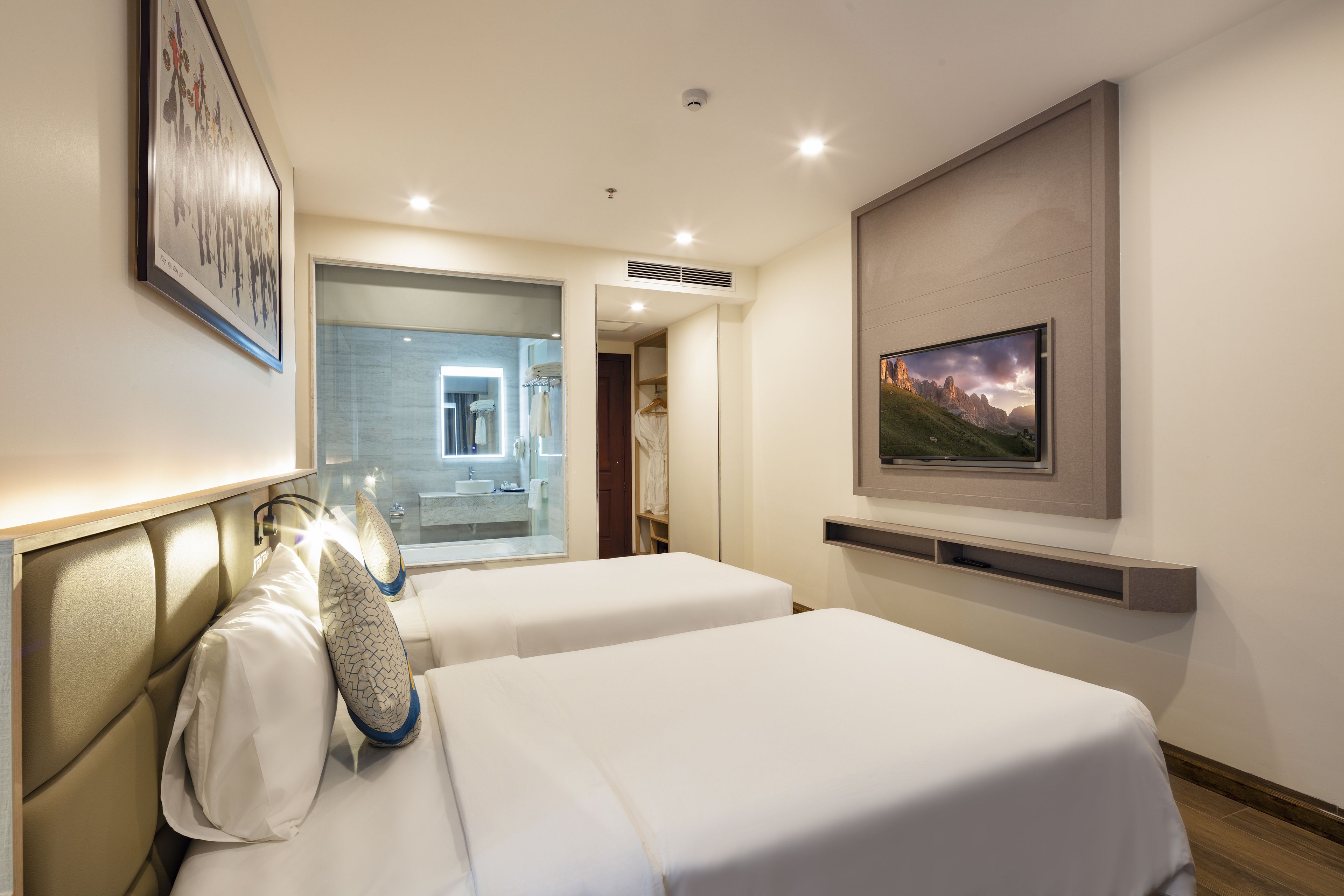 Ein Hotelzimmer mit zwei Betten, einem durch eine Glastür sichtbaren Badezimmer und einem an der Wand montierten Fernseher.