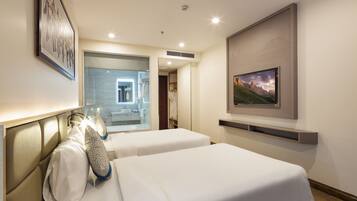 Ein Hotelzimmer mit zwei Betten, einem durch eine Glastür sichtbaren Badezimmer und einem an der Wand montierten Fernseher.