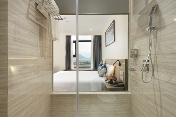 A modern hotel bathroom with a glass shower enclosure, a bed with white linens, a bedside table with a phone and a vase of flowers, and a view of a cityscape through the window. - Lucky Sun Hotel (Nha Trang)