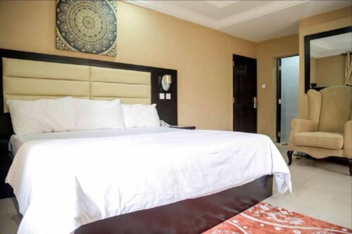 A hotel room with a large bed, a headboard, a wall-mounted artwork, and a seating area with a chair.