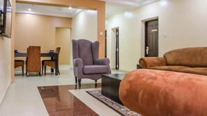 A living room with a sofa, armchair, and a coffee table. There is a dining area with chairs and a table in the background. - The Capital Apartments (Abuja)