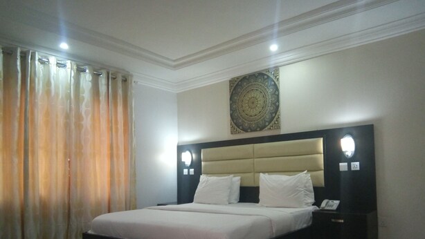 A bedroom with a large bed, a headboard with built-in lighting, a bedside table with a phone, and a decorative wall piece. - The Capital Apartments (Abuja)