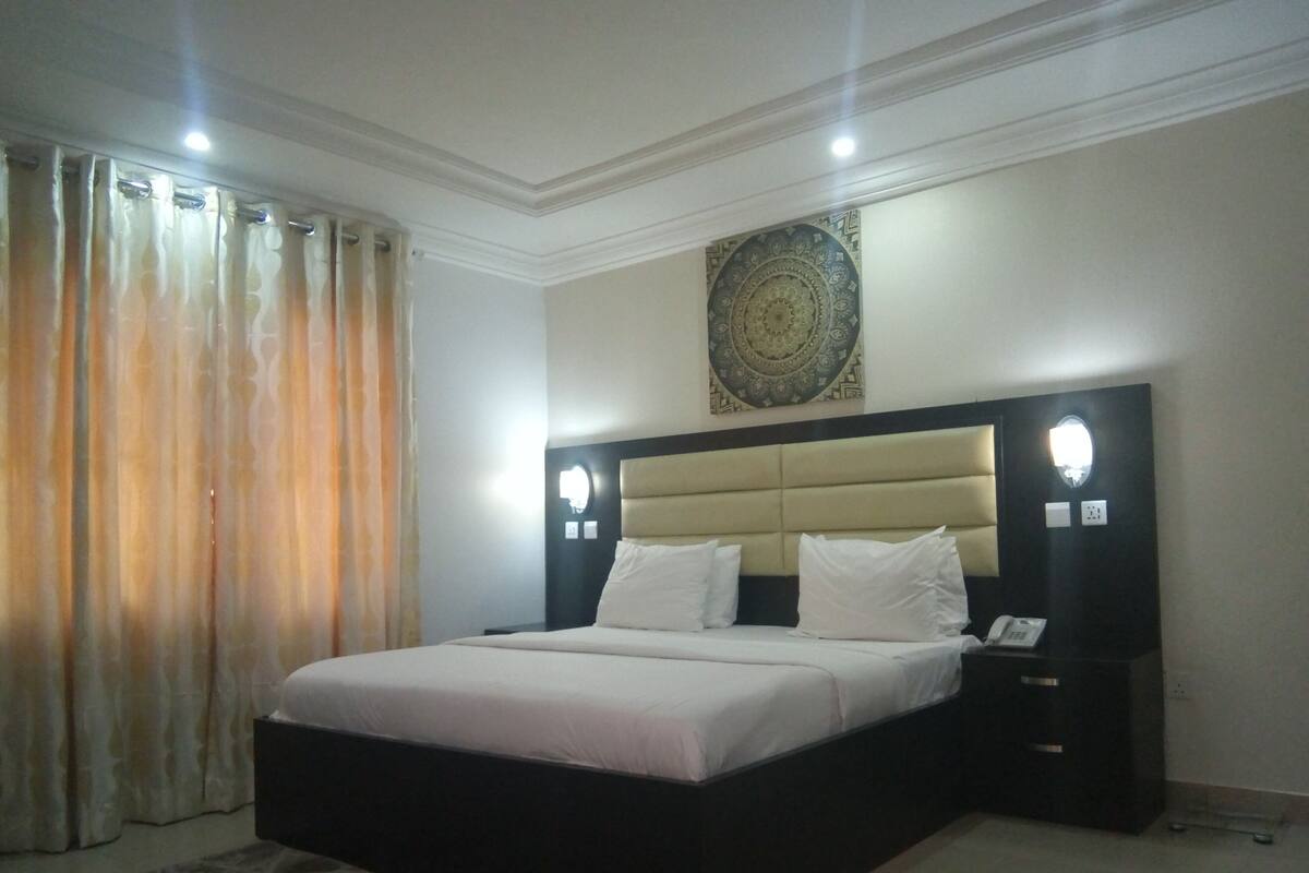 A bedroom with a large bed, a headboard with built-in lighting, a bedside table with a phone, and a decorative wall piece.
