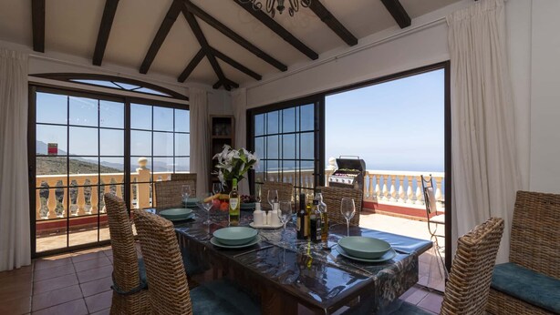 Ein Essbereich mit Tisch für vier Personen, großen Fenstern, Korbstühlen und Blick auf das Meer.