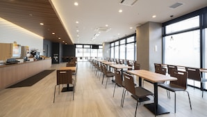 A modern cafeteria with long tables and chairs, a counter with a coffee machine, and large windows allowing natural light. - Washington R&B Hotel Nagoya Shinkansenguchi (Nagoya)