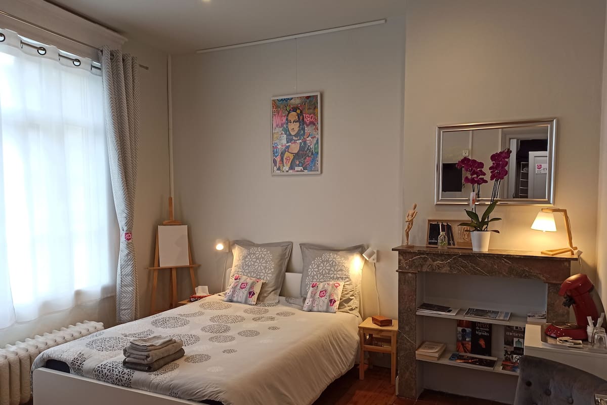 Un dormitorio ordenado con cama, mesitas de noche, un escritorio pequeño, un espejo y una chimenea.