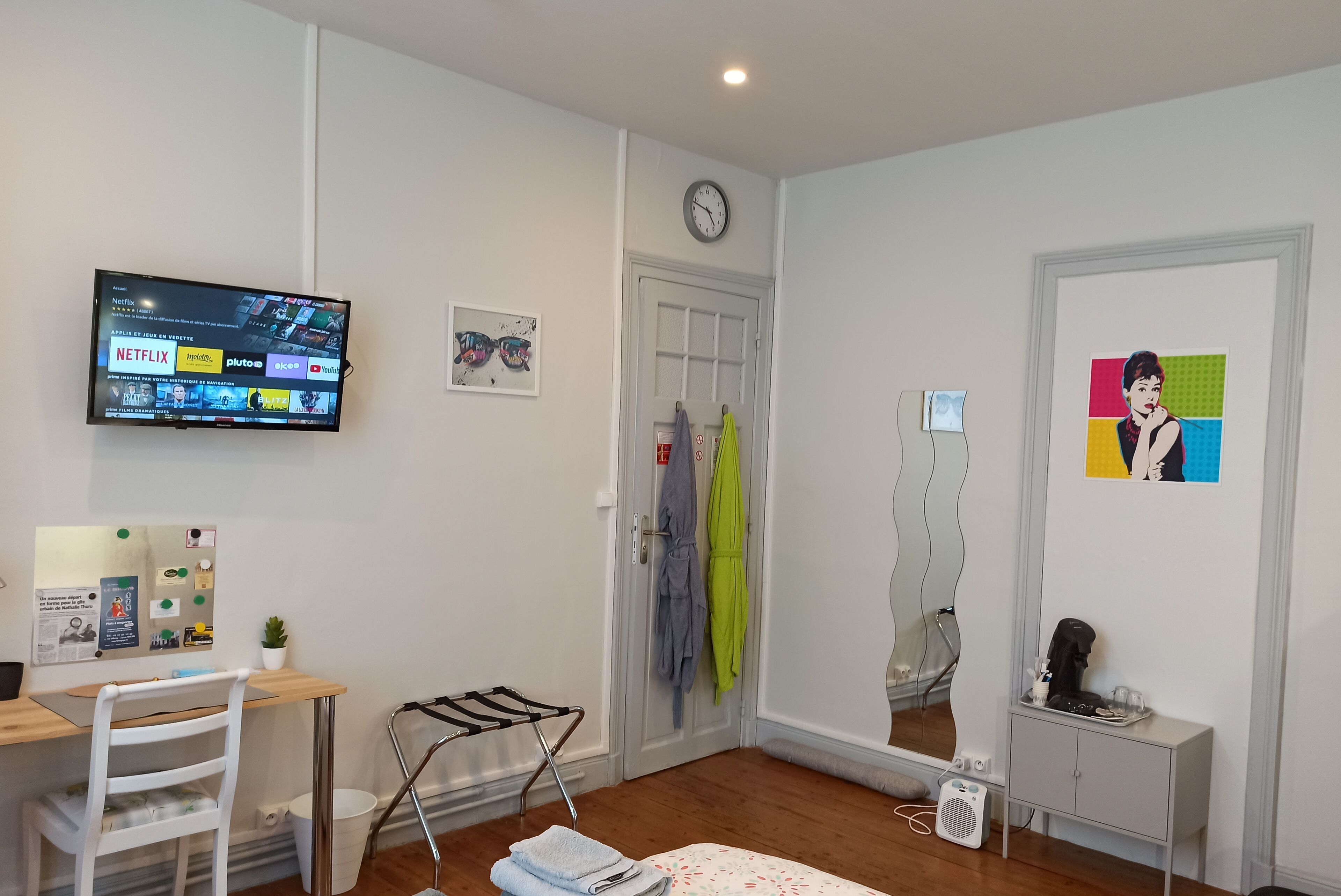 Ett modernt vardagsrum med en platt-TV, ett skrivbord i trä, en vit stol, en liten krukväxt, en väggklocka och ett inramat konstverk.