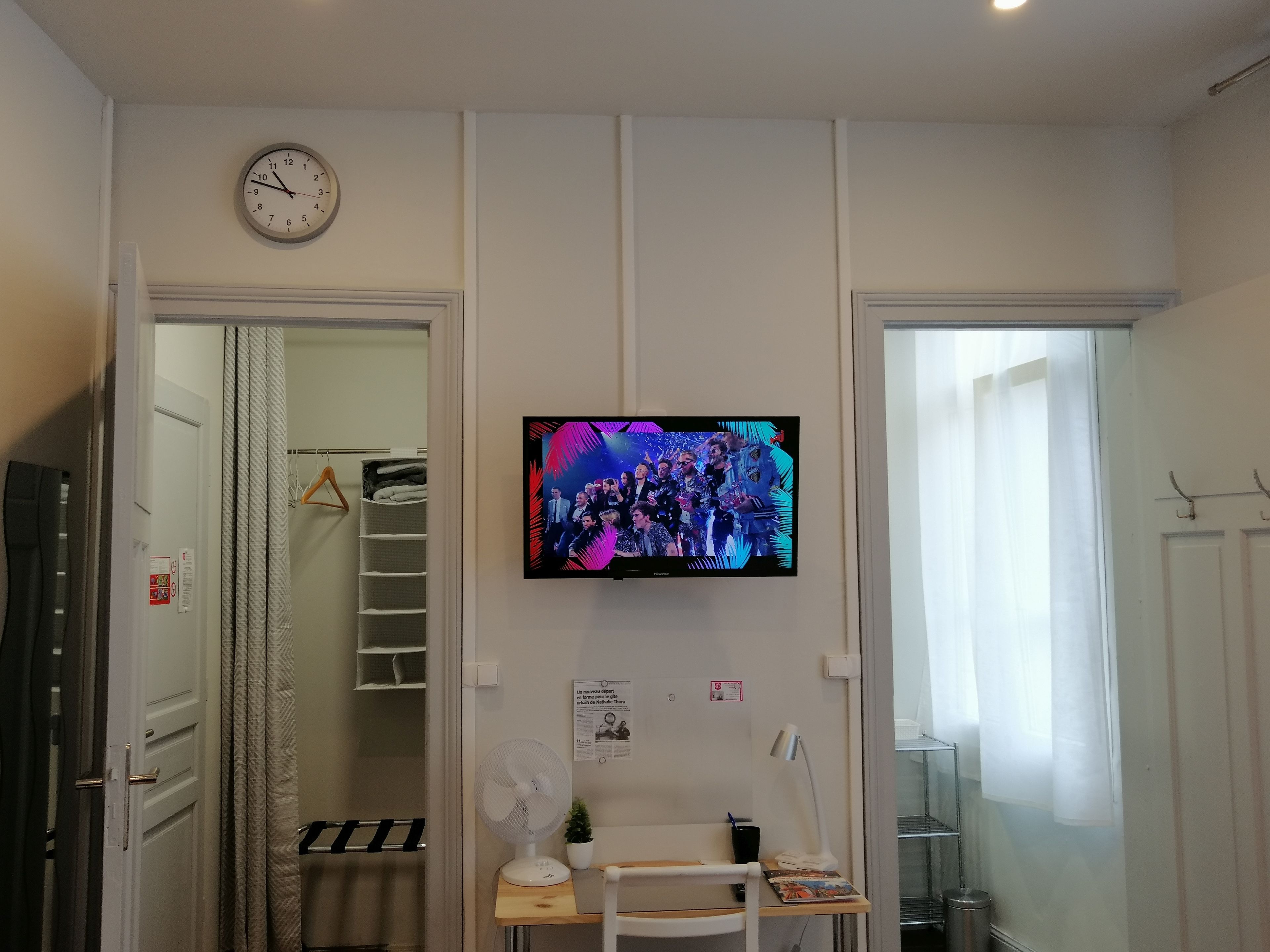 A hotel room with a television mounted on the wall, a clock, a wardrobe, a desk with a chair, and a window with curtains.