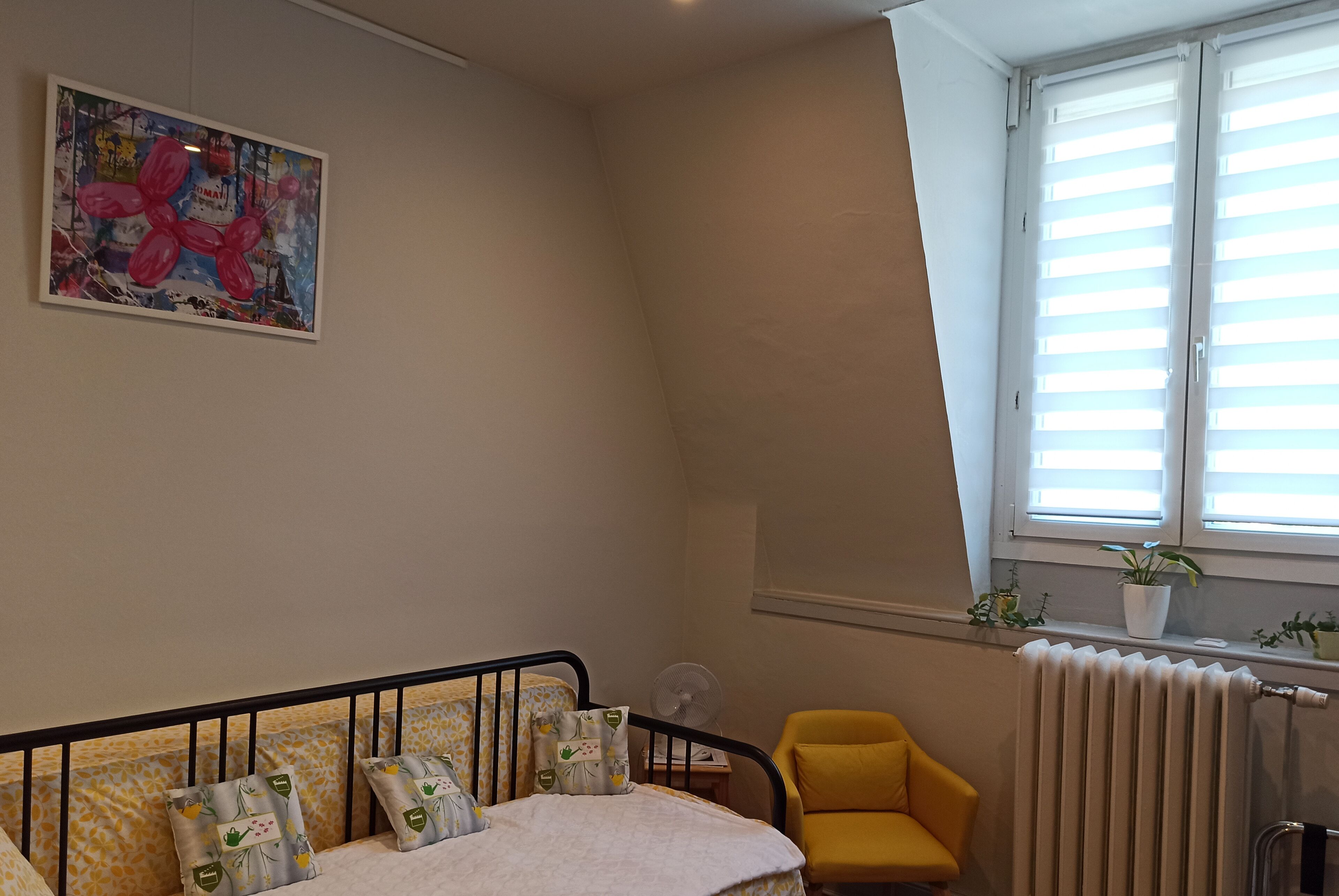 Een slaapkamer met een bed, een gele stoel, een radiator en een raam met zonwering.