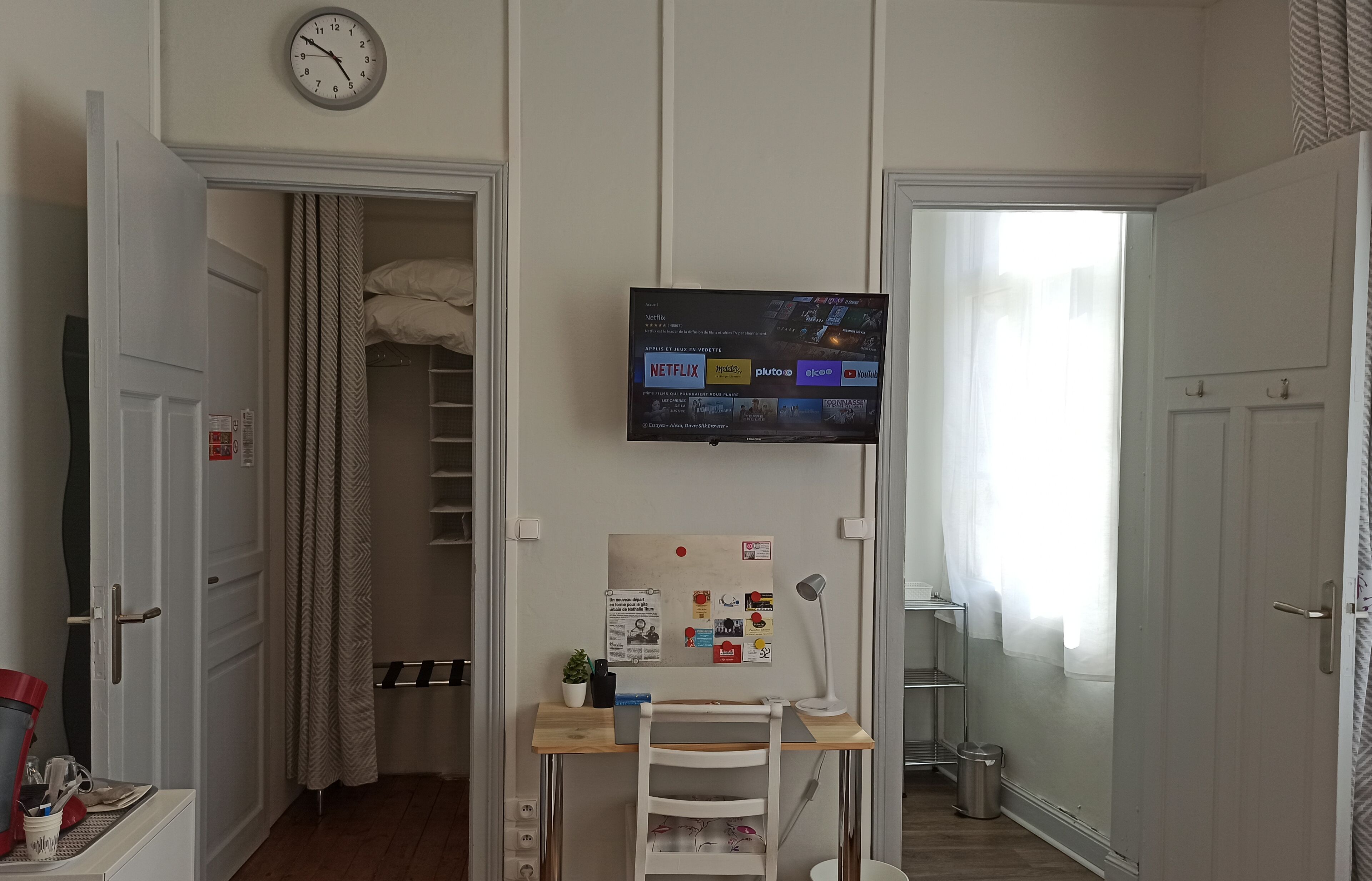 A bedroom with a bed, a desk with a chair, a TV mounted on the wall, and a window with curtains.