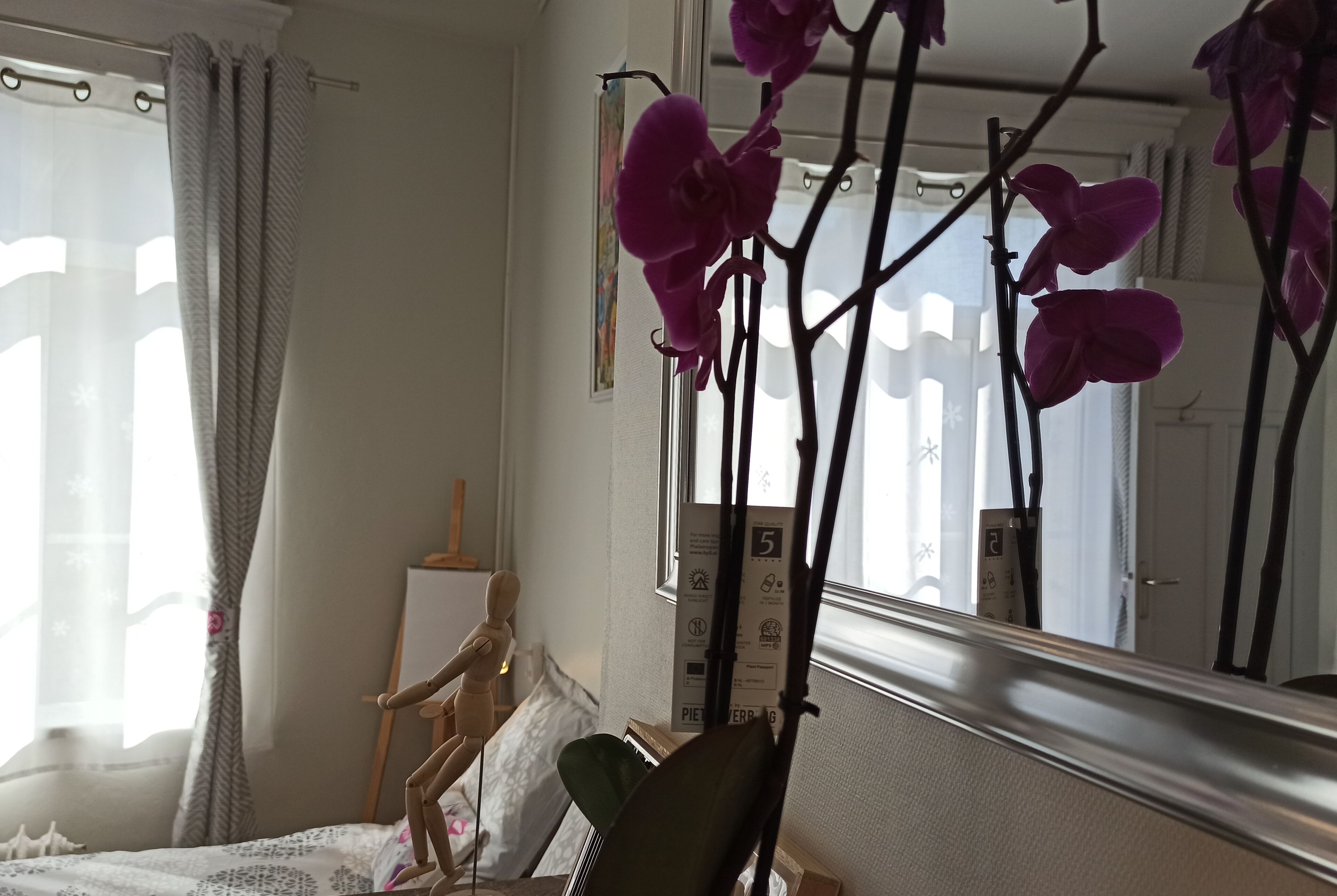 Un dormitorio con una cama, un maniquí de madera, una silla y un espejo que refleja orquídeas.