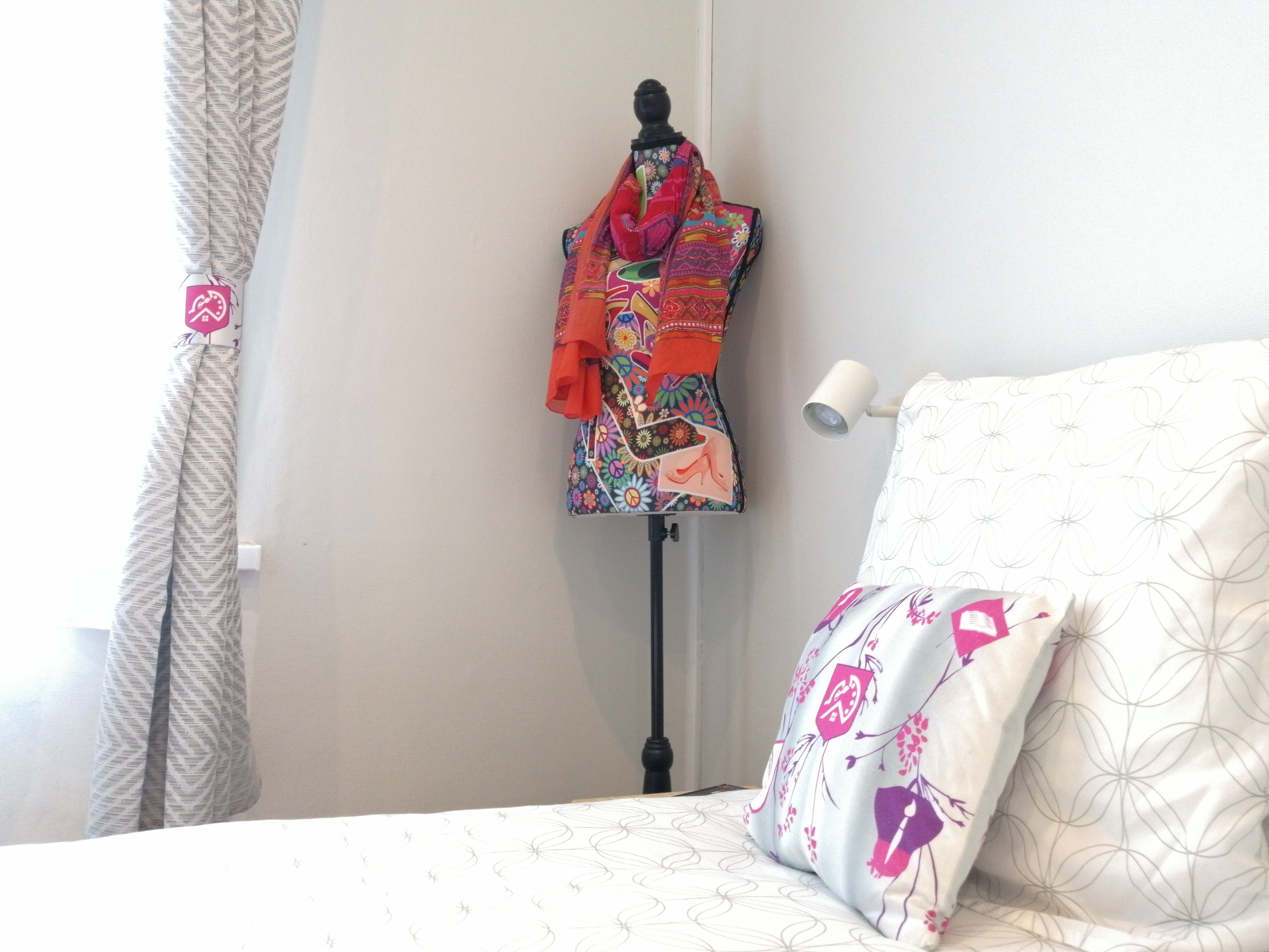 A bedroom with a bed, a mannequin dressed in colorful clothing, a lamp, and patterned curtains.