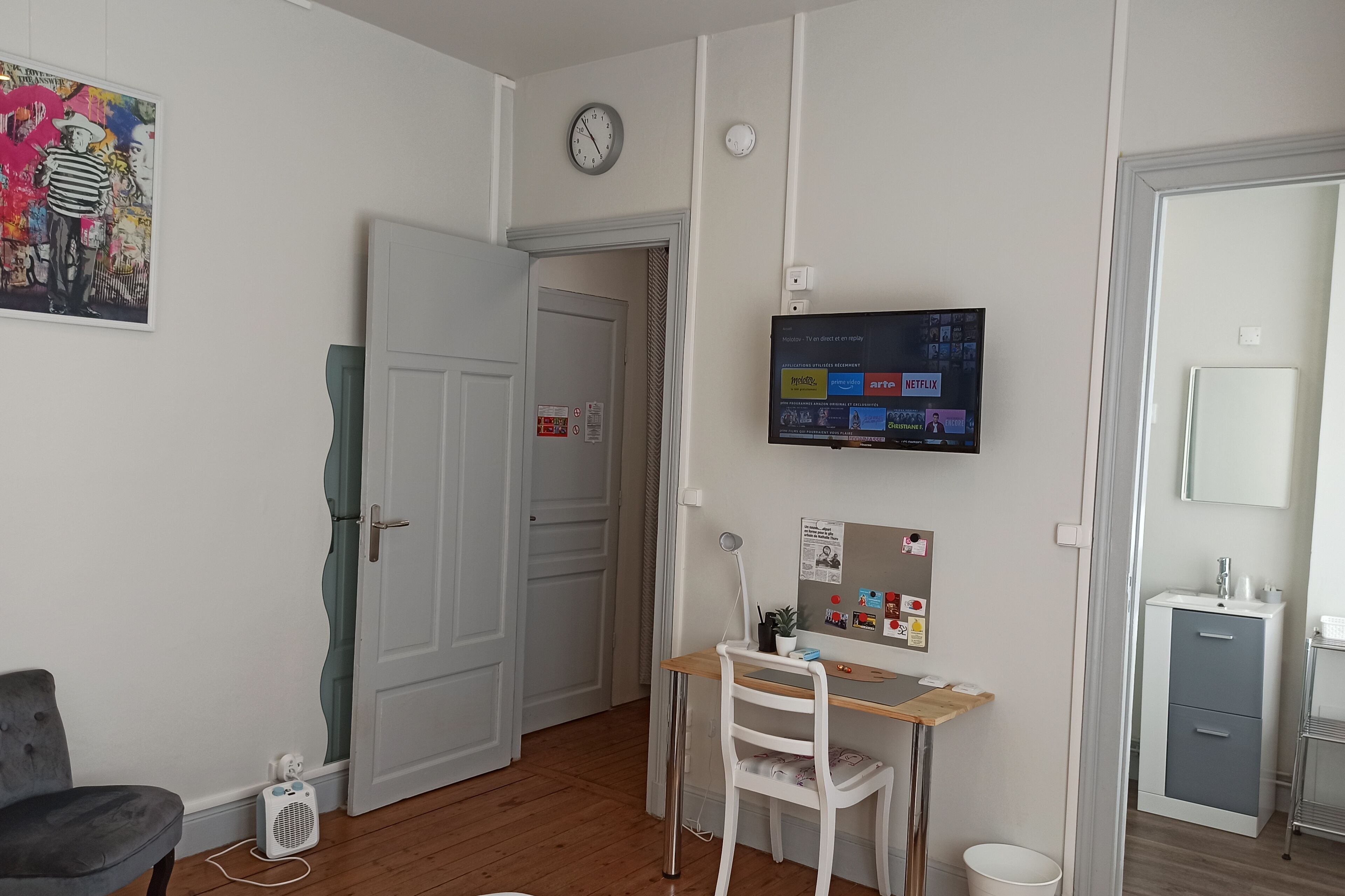 Una habitación con un escritorio de madera, una silla, un televisor montado en la pared, un reloj y una puerta.