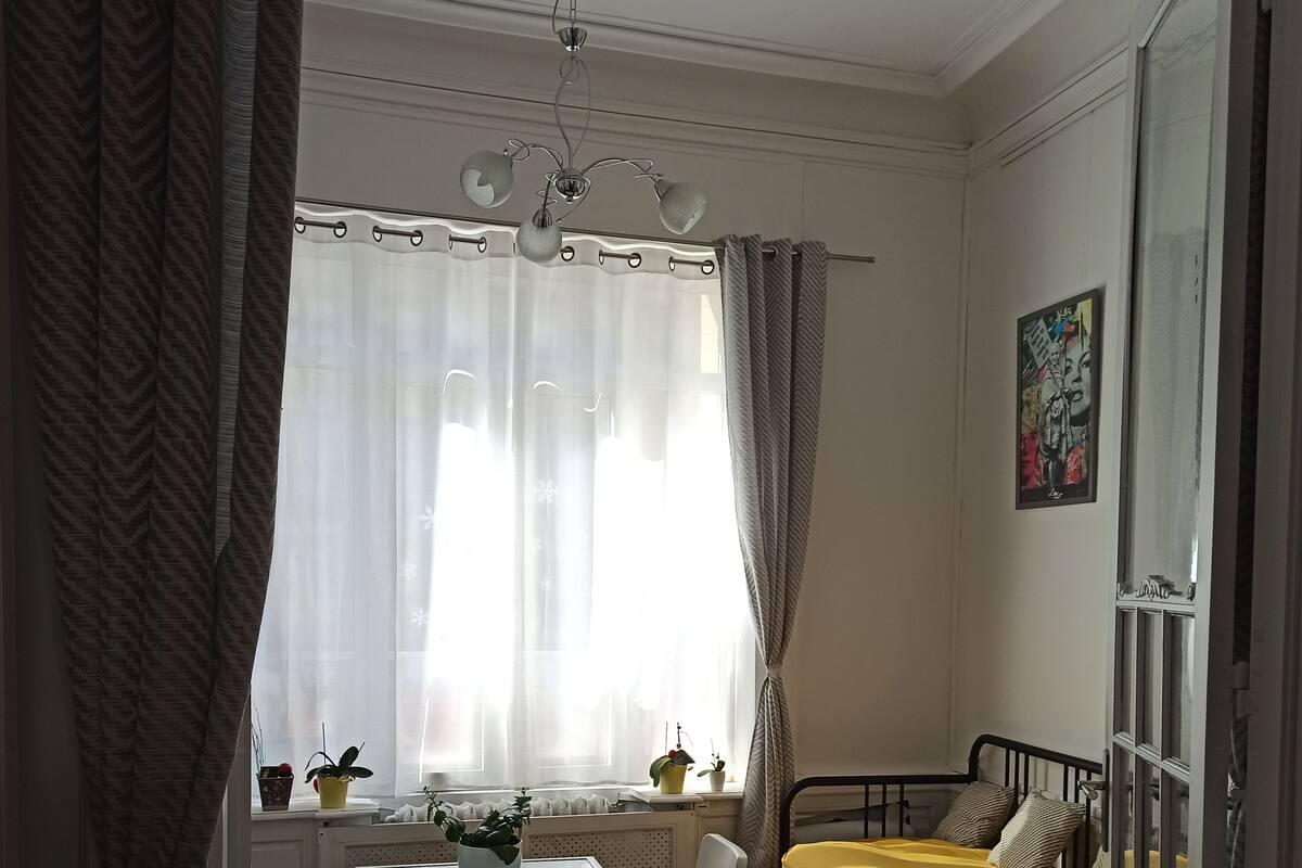 Una habitación con cama, una mesa con sillas, un ventanal con cortinas y una lámpara en el techo.