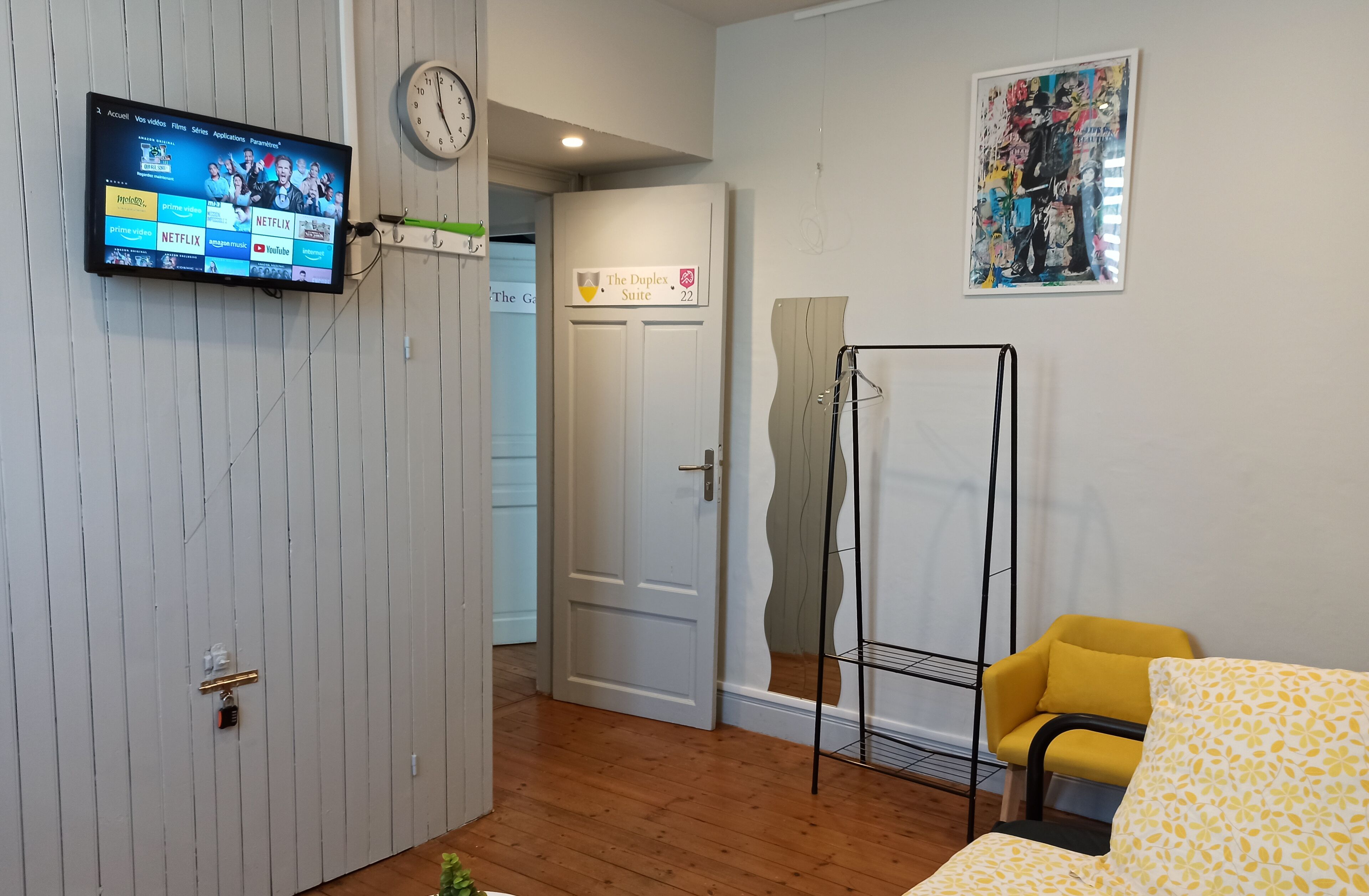 Una habitación de hotel con un televisor montado que muestra servicios de transmisión, una puerta con la etiqueta "The Duplex Suite 22", una silla amarilla y un estante fijado a la pared.