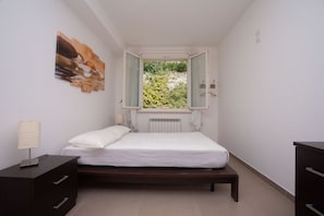 A bedroom with a bed, a dresser, a nightstand, a window with a view of greenery, and a painting on the wall.