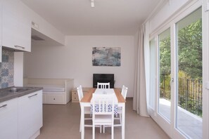 A modern kitchen with white cabinets, a dining table with chairs, a flat-screen TV, and a balcony with a view of trees.