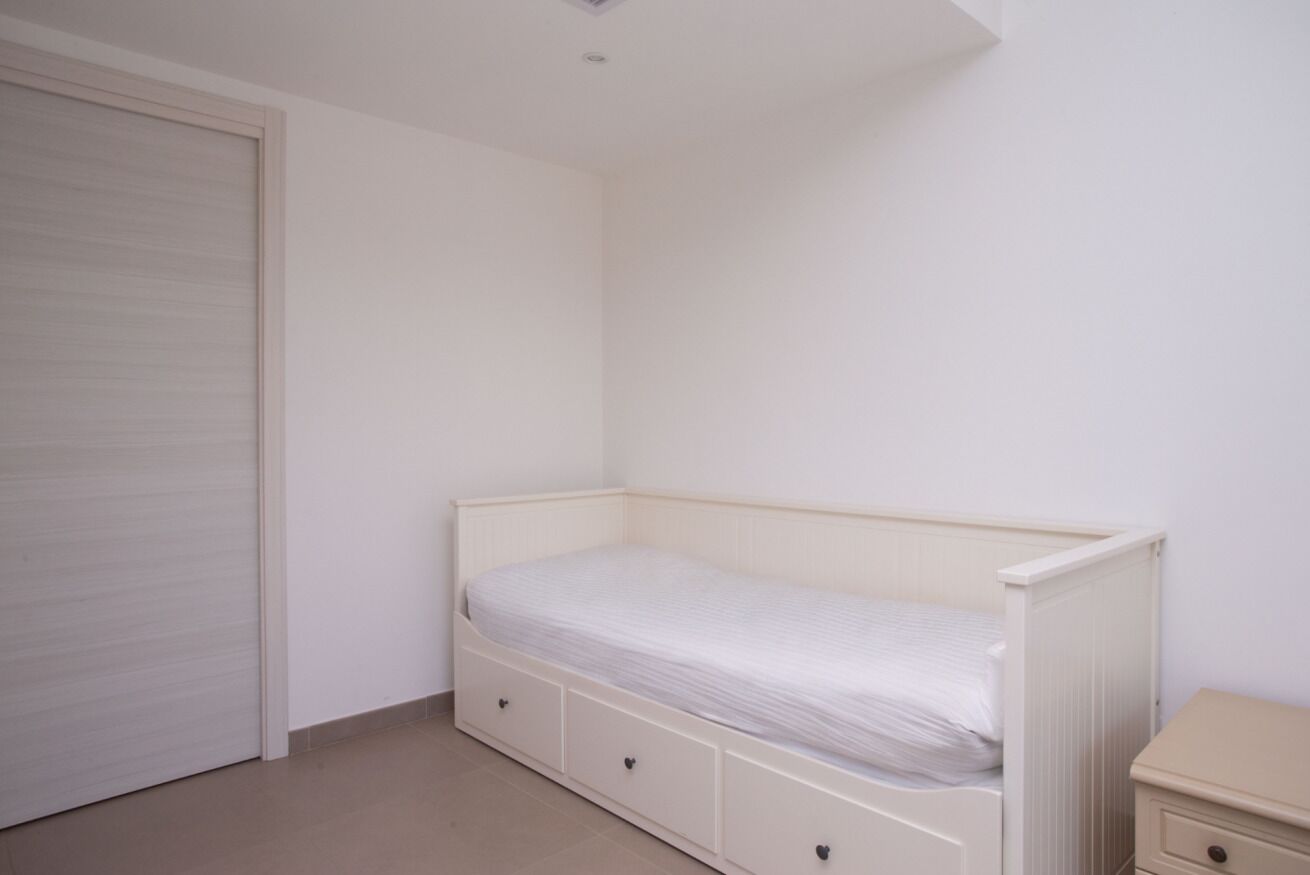Een kleine kamer met een enkel bed, een gesloten deur, een nachtkastje en een ladekast.