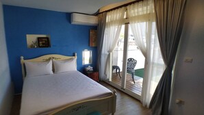 Quarto com cama, ar condicionado, mesa de cabeceira, espelho e vista para o exterior através de uma janela com cortinas.