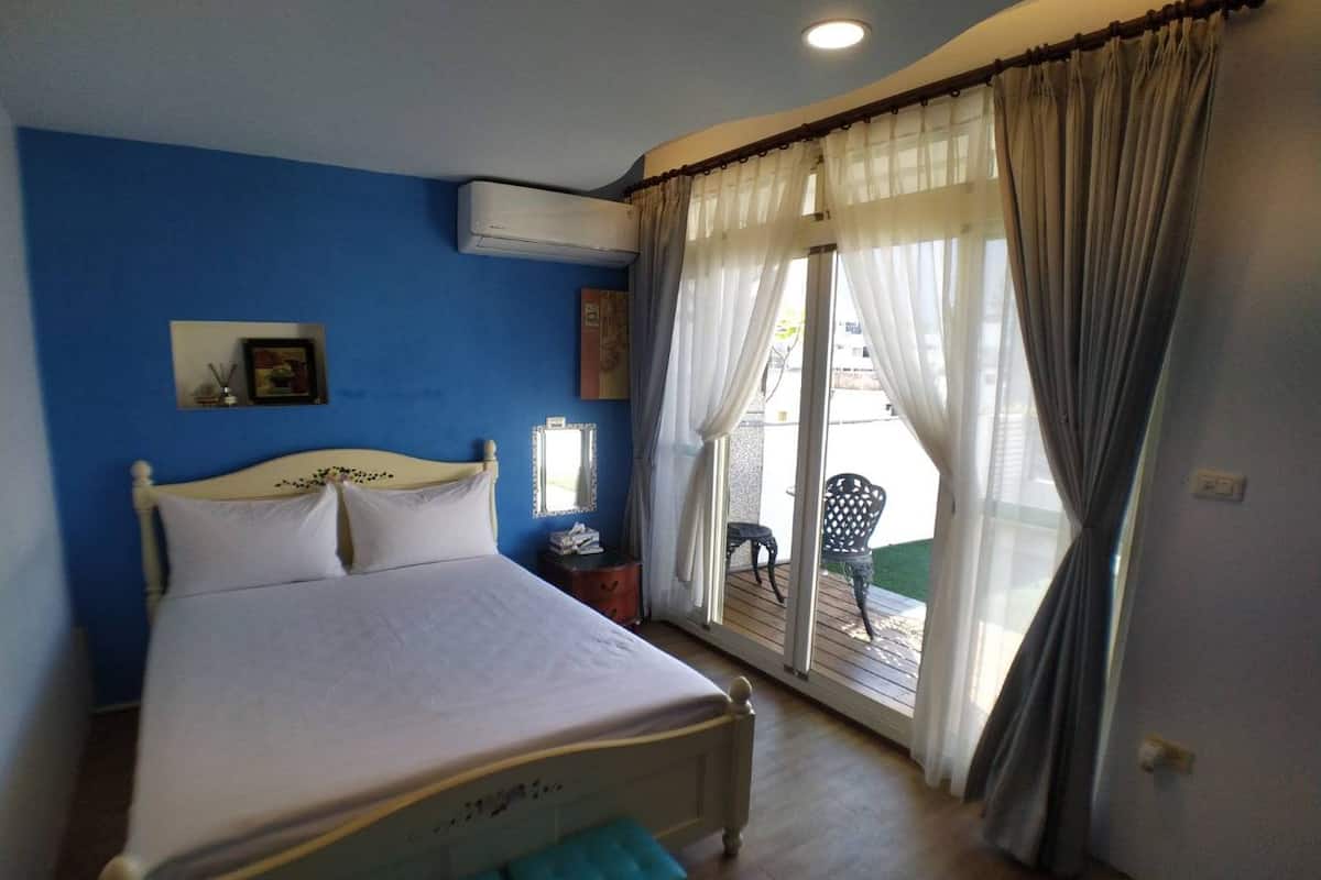 Un dormitorio con cama, unidad de aire acondicionado, mesita de noche, espejo y vistas al exterior a través de una ventana con cortinas.