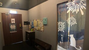 Um corredor com uma porta de madeira, um mural de avisos, um pequeno banco de madeira e uma cortina decorativa com motivos de gato e flores.