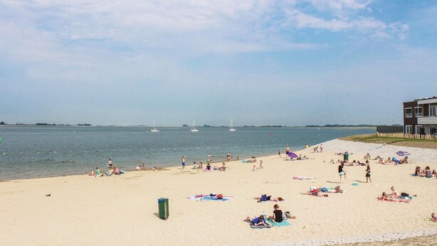 A beach with people sunbathing and swimming, a building on the right, and sailboats in the distance.