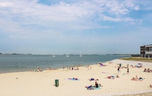 Una playa con gente tomando el sol y nadando, un edificio a la derecha y veleros a lo lejos.