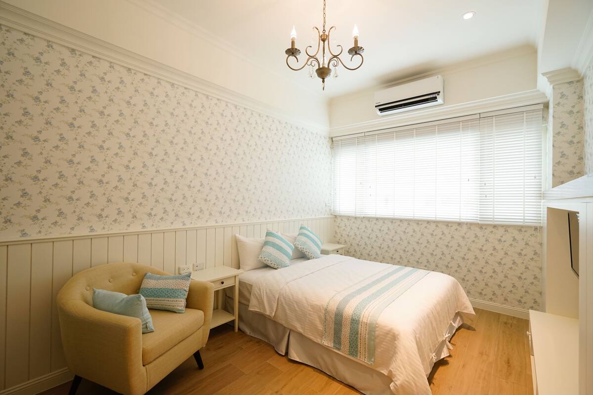 A bedroom with a bed, a chair, a nightstand, a chandelier, and an air conditioning unit.