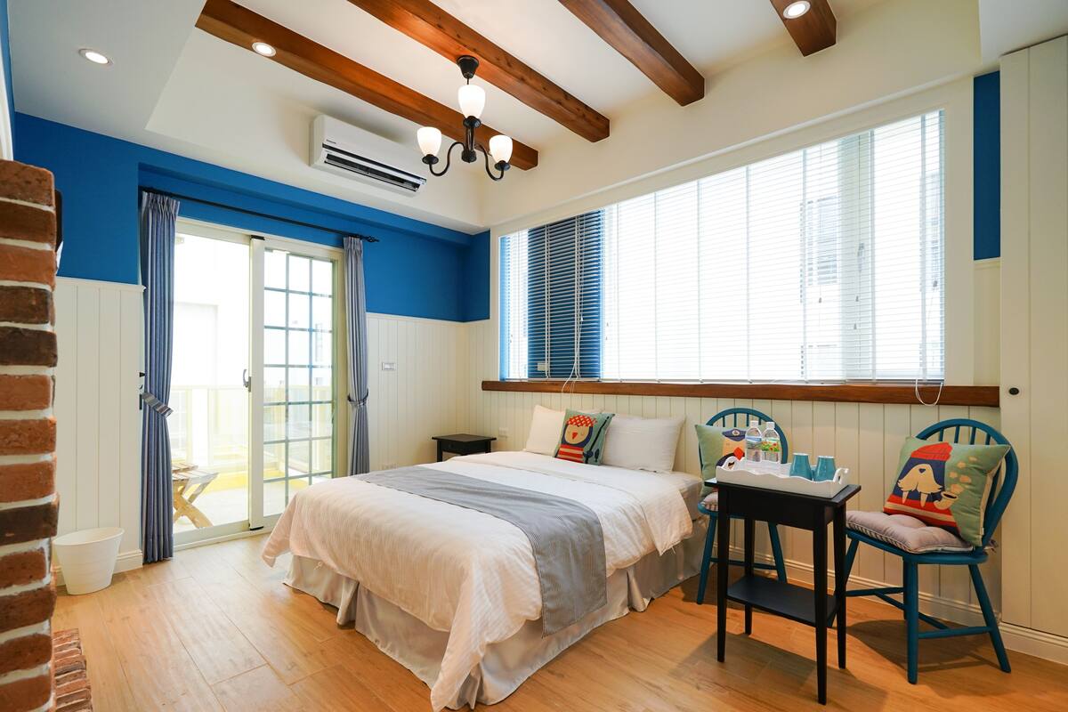 A bedroom with a large bed, a wooden floor, a window with blinds, a table with a chair, and a brick wall.