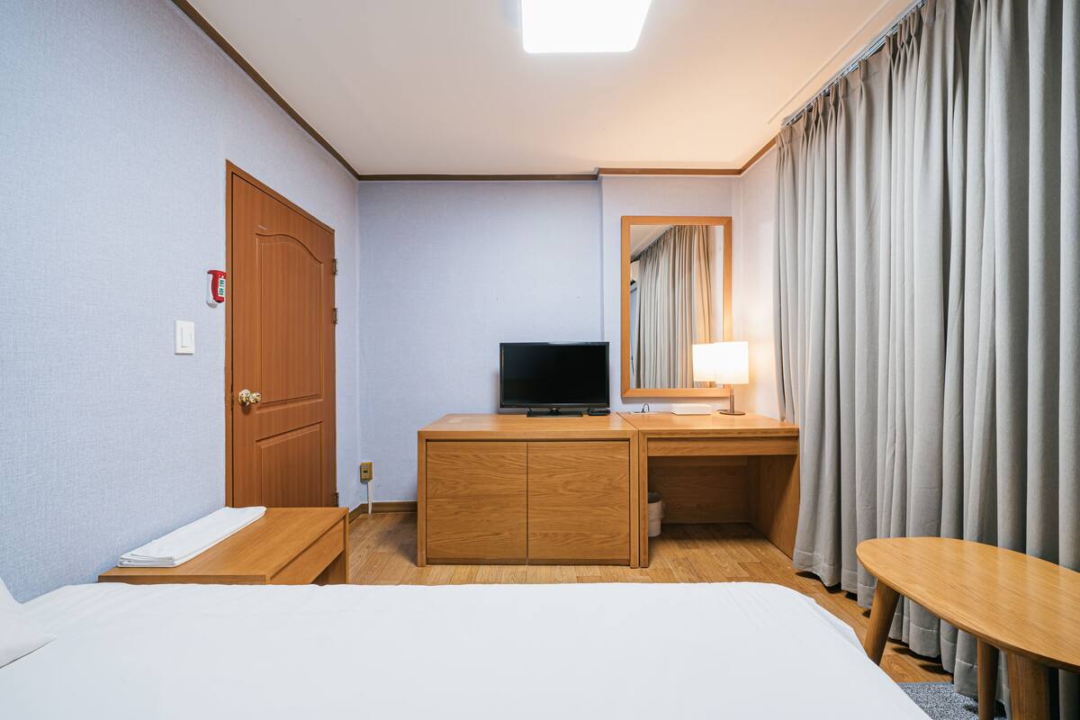 Une chambre d’hôtel avec un lit, un bureau en bois, une télévision, un miroir et une fenêtre avec des rideaux.