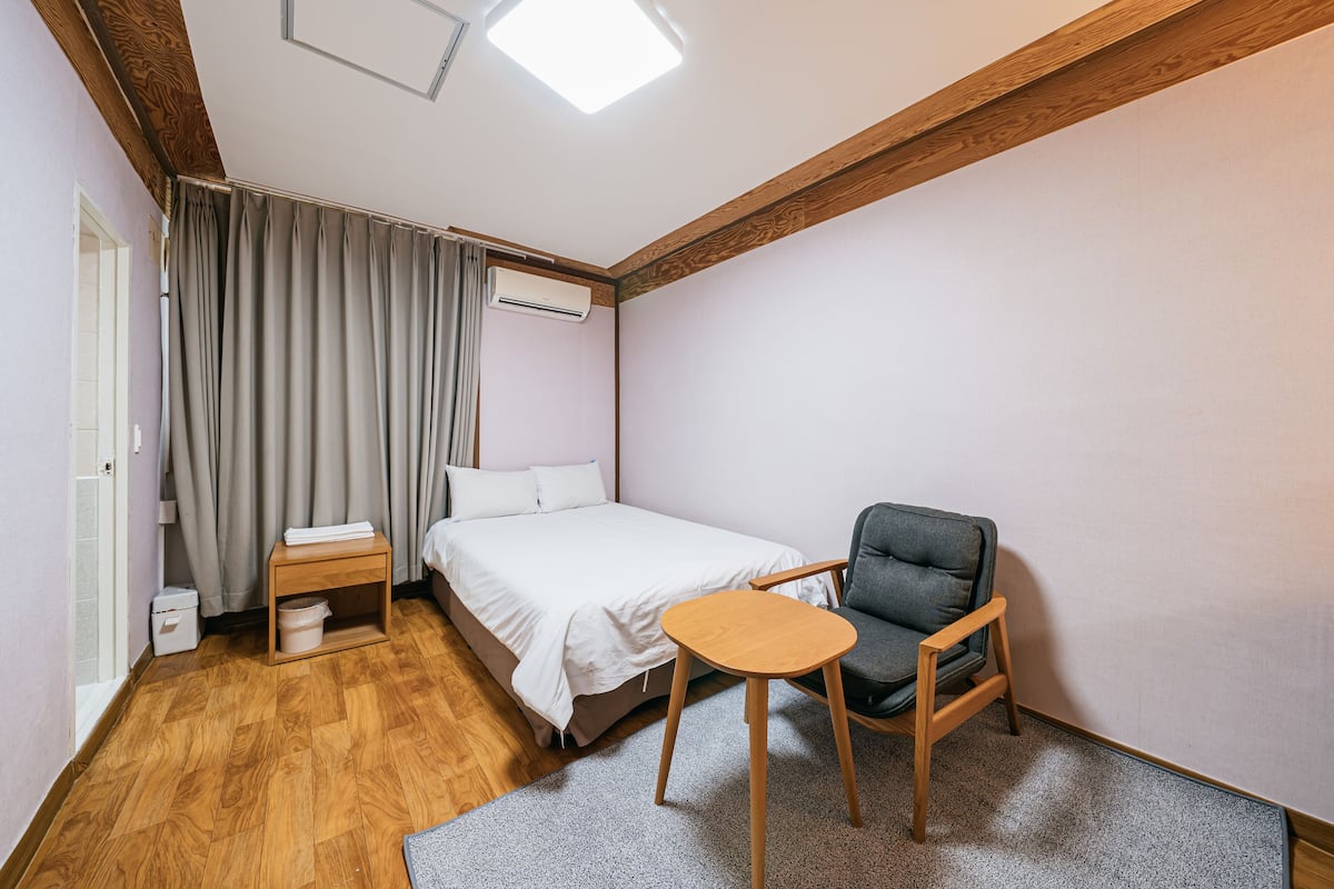 Une petite pièce bien éclairée, comprenant un lit, une chaise, une petite table et un appareil de climatisation.