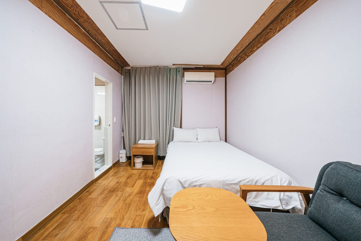 Une petite chambre bien éclairée, avec un lit simple, une table de chevet, une chaise et une fenêtre avec des rideaux.