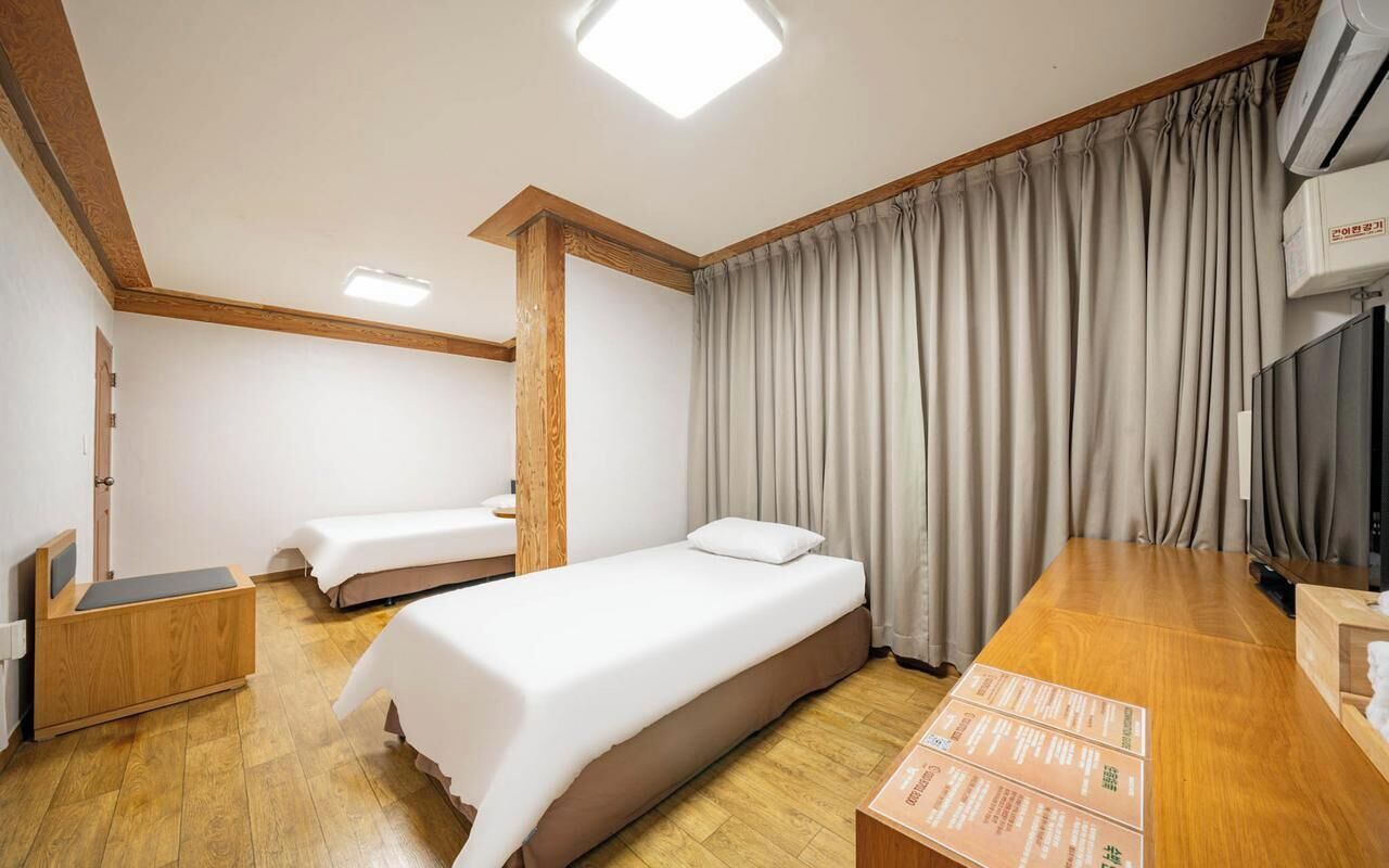 Une chambre d’hôtel avec deux lits simples, un bureau en bois, une télévision et une grande fenêtre avec des rideaux.