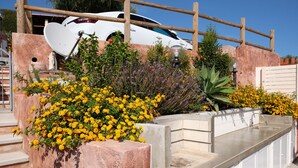 A white car parked behind a wooden fence, with a concrete planter filled with yellow flowers and a small garden area. - Villa Cinzia sea view (Porto Tramatzu)