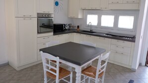 A kitchen with white cabinets, a black countertop, a built-in oven, a microwave, and a sink. There is a window with blinds and a white chair with a woven seat. - Villa Cinzia sea view (Porto Tramatzu)