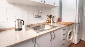 A kitchen with a stainless steel sink, a kettle, a coffee maker, a bottle of oil, a can of soda, a cutting board, and a washing machine. - 2 bedroom beautiful apartment in Pineda de Mar (Pineda de Mar)