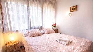 A neatly made bed with a patterned bedspread and pillows, a bedside lamp, a wicker side table, a framed picture on the wall, and a potted plant with red flowers. - 2 bedroom beautiful apartment in Pineda de Mar (Pineda de Mar)