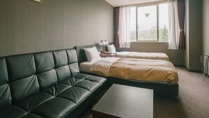 Habitación de hotel con sofá de cuero oscuro, dos camas individuales, una mesita y una ventana con cortinas.