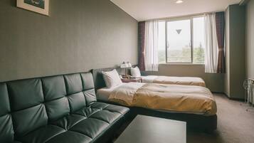 Habitación de hotel con sofá de cuero oscuro, dos camas individuales, una mesita y una ventana con cortinas.