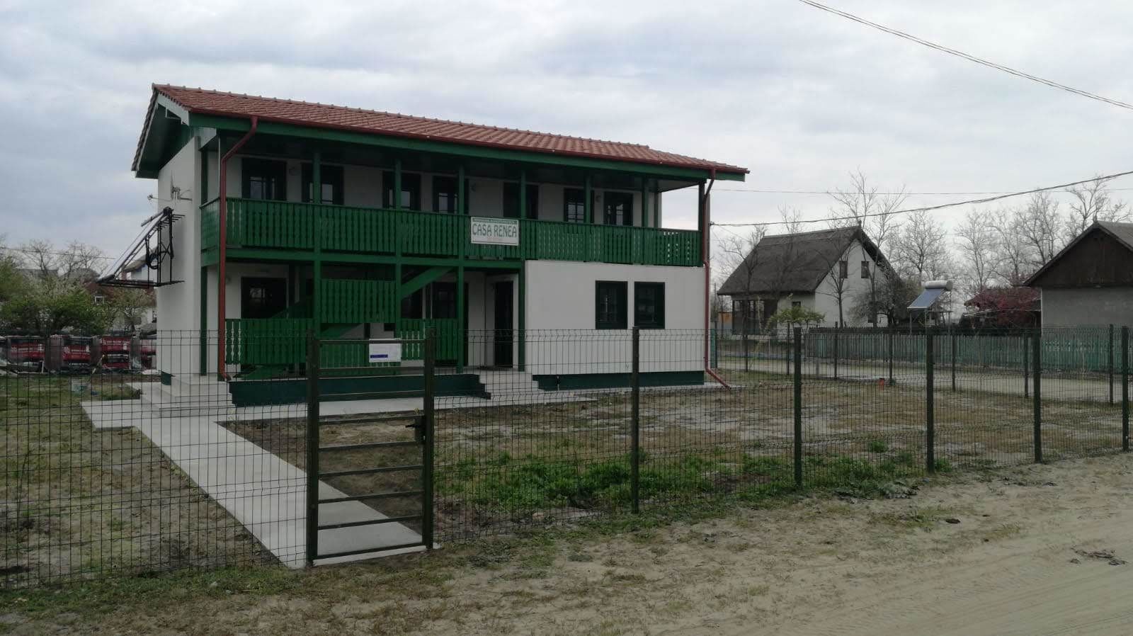 Een gebouw met twee verdiepingen, een groen-witte gevel, een overdekte veranda en een omheinde tuin.