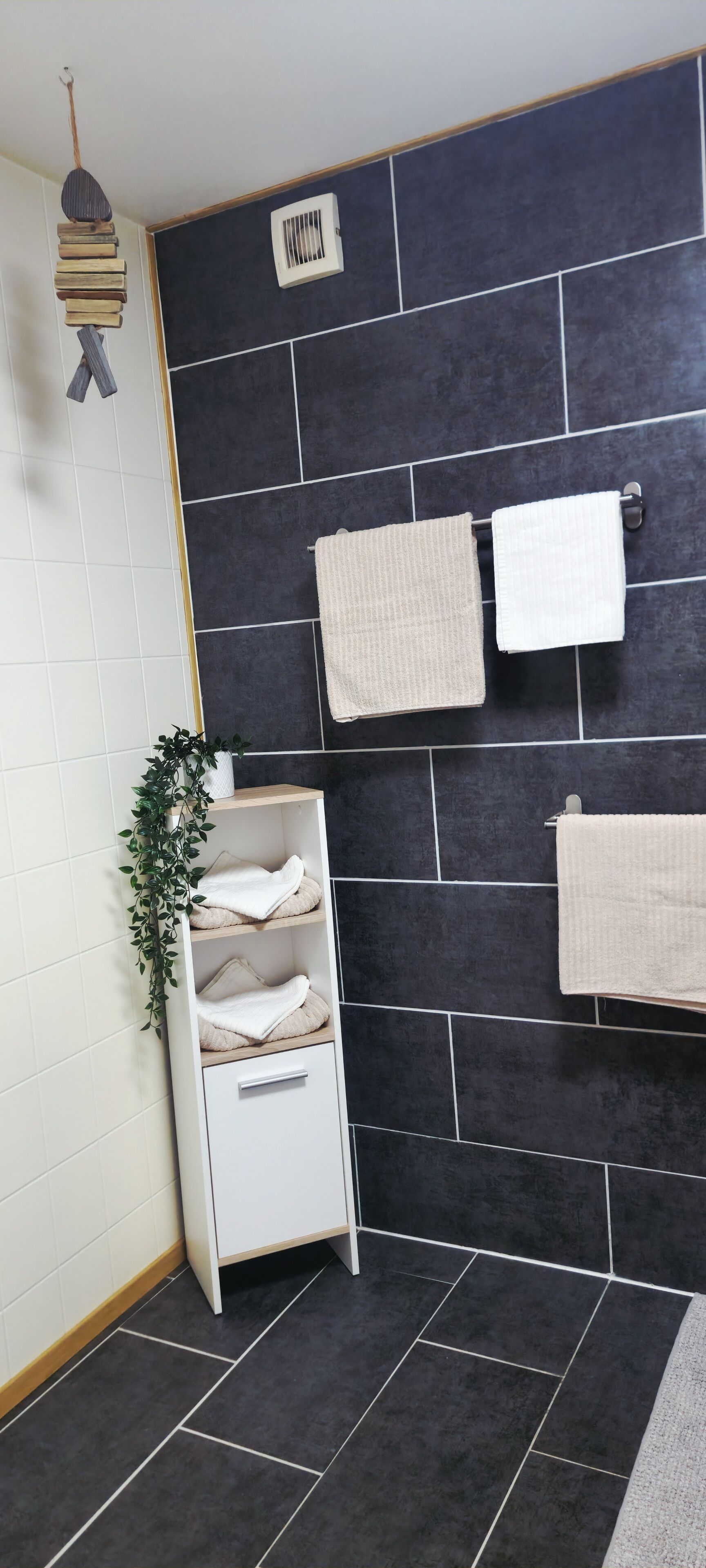 Un baño con paredes de baldosas oscuras, dos toallas colgadas en un perchero, un estante blanco con toallas dobladas y una planta colgante.