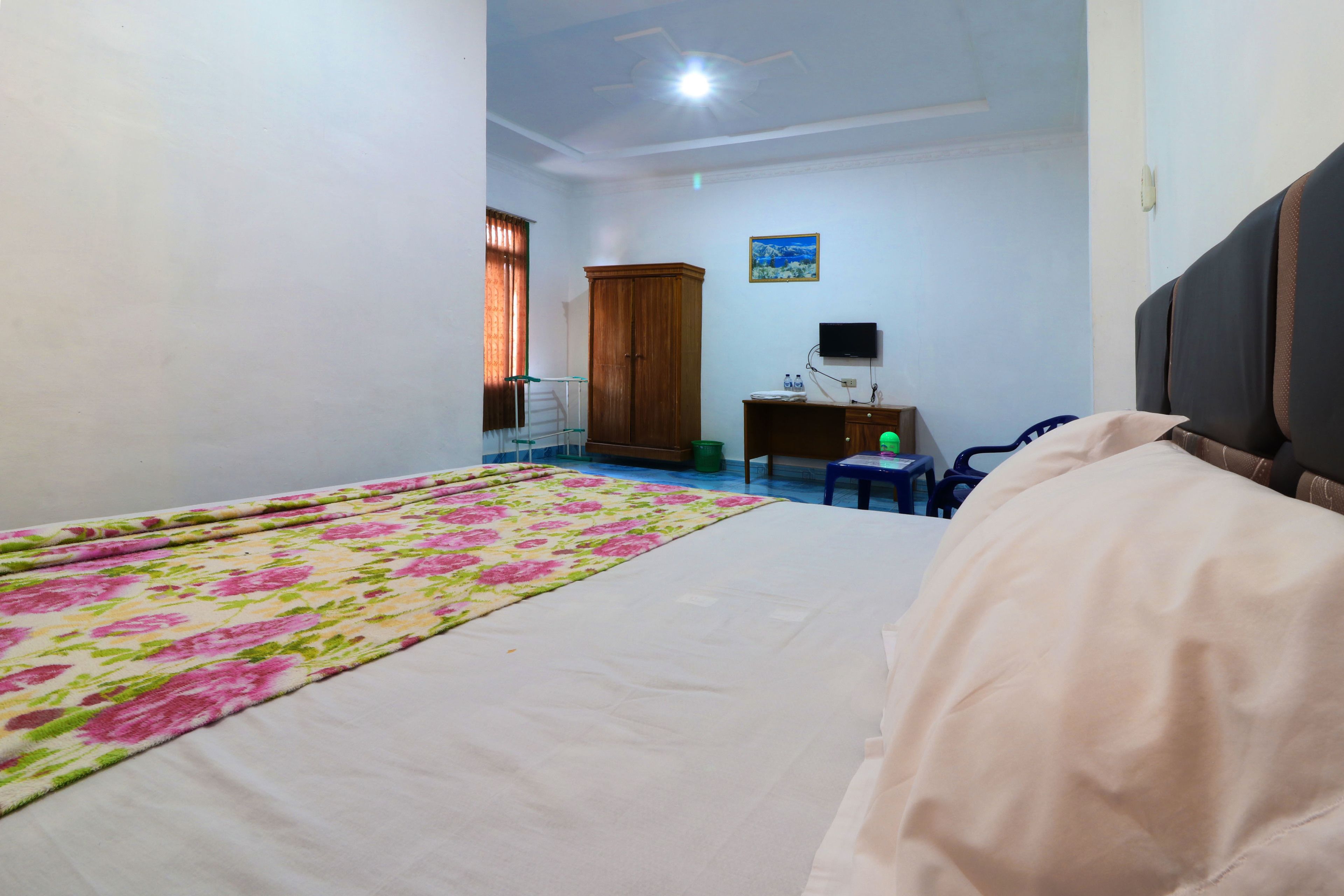 A bedroom with a bed, a wooden wardrobe, a desk with a chair, and a television.