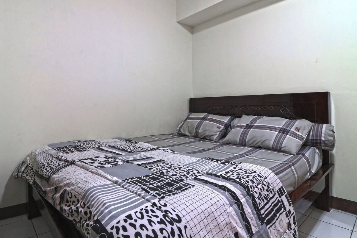 A bed with a patterned bedspread and pillows, a wooden headboard, and a plain wall.