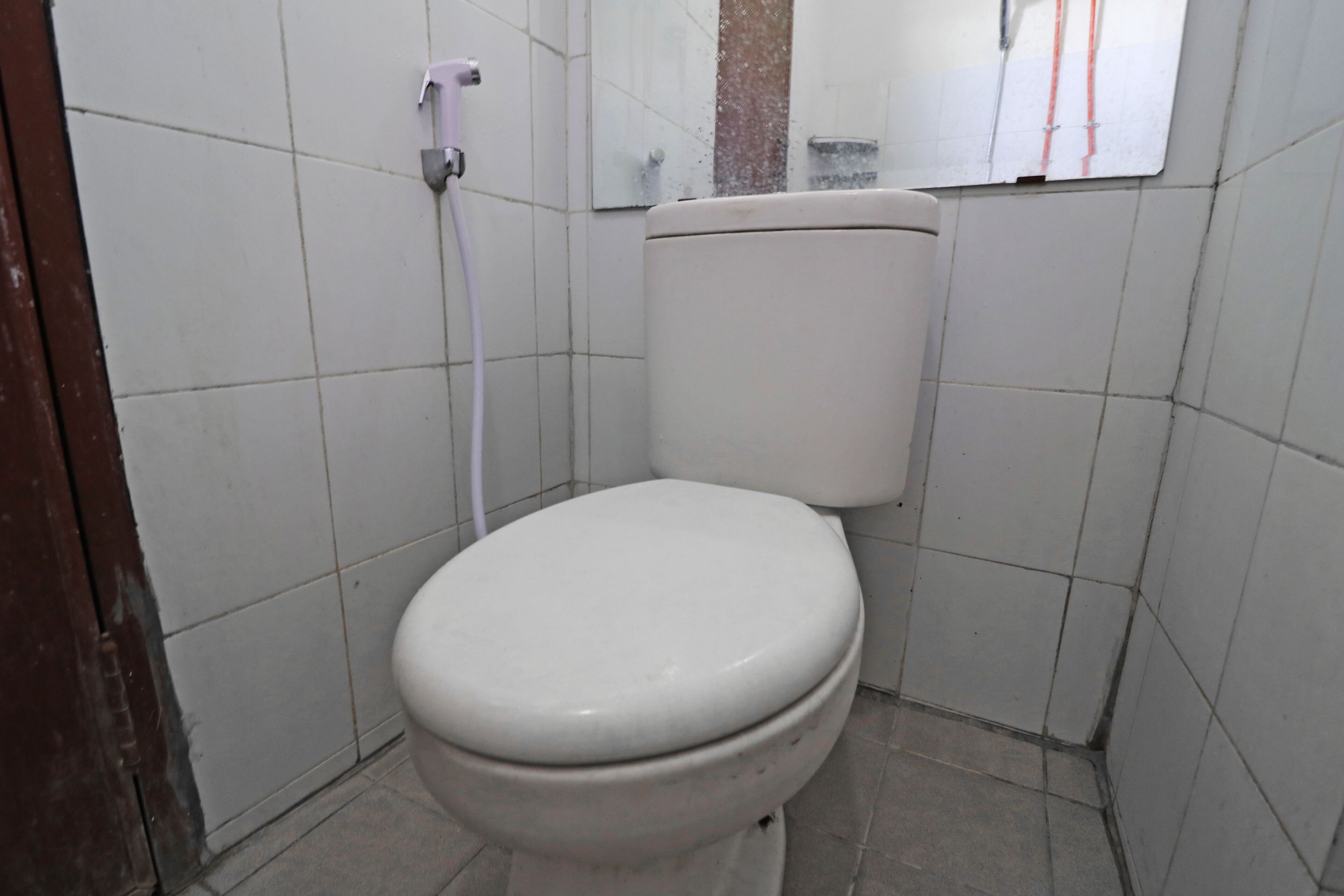 A standard white toilet in a bathroom with white tiles, a visible pipe, and a mirror above the toilet.