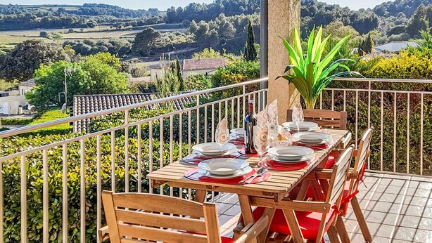 An outdoor dining area with a table set for four, surrounded by wooden chairs, overlooking a scenic landscape with hills and trees.